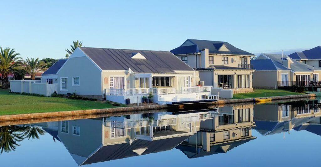 Luxury modern homes reflecting on a peaceful, clear lake during daylight, showcasing architectural beauty.